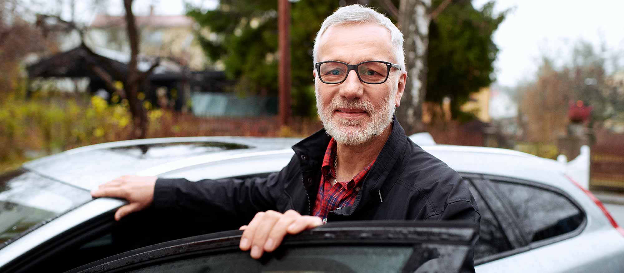 Äldre man med skägg och glasögon står vid framdörren till sin bil