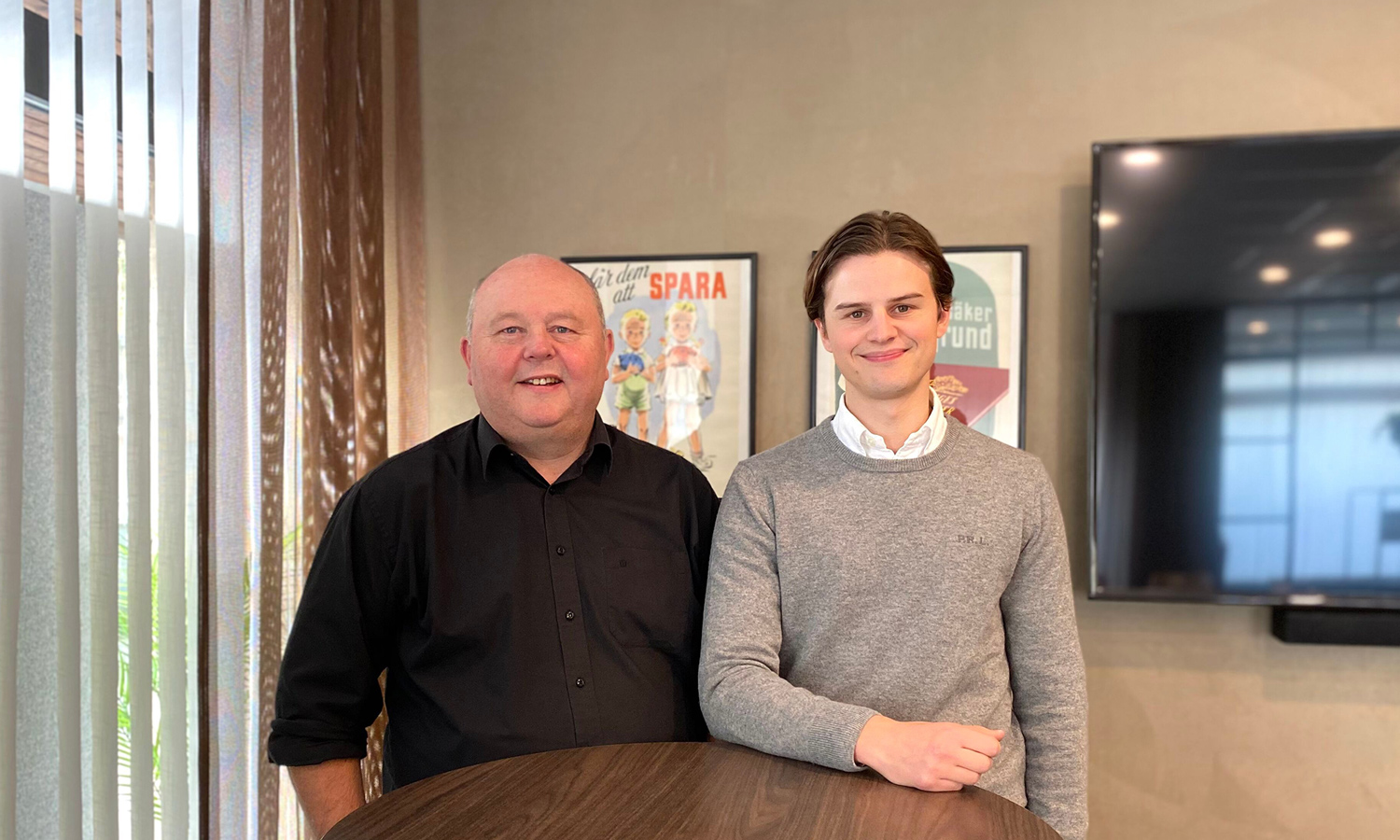 Roger and Axel are standing by a high table, smiling at the camera. They are in a conference room.