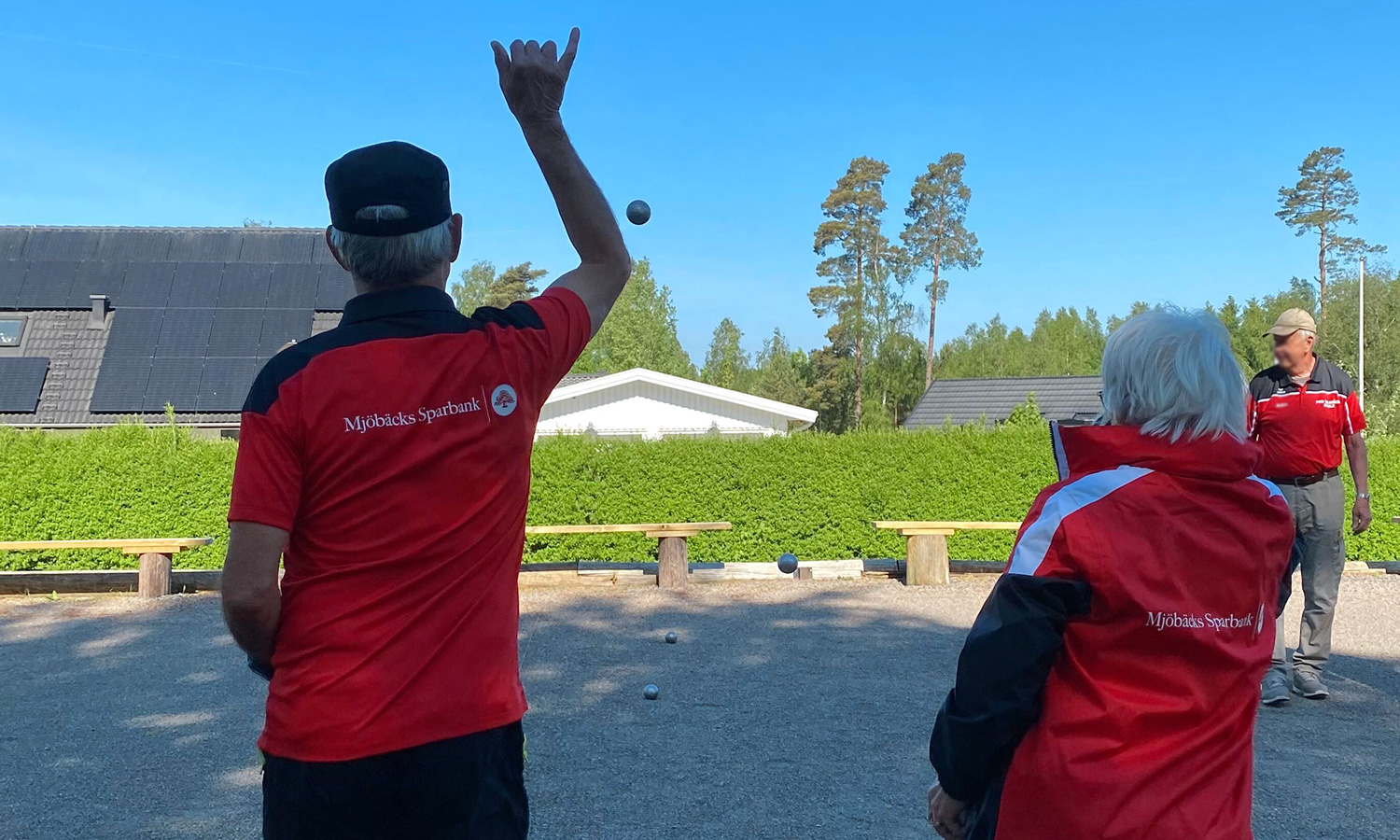 Two pensioners play boule. Wearing match kit with Mjöbäcks Sparbank's logo printed on the back.